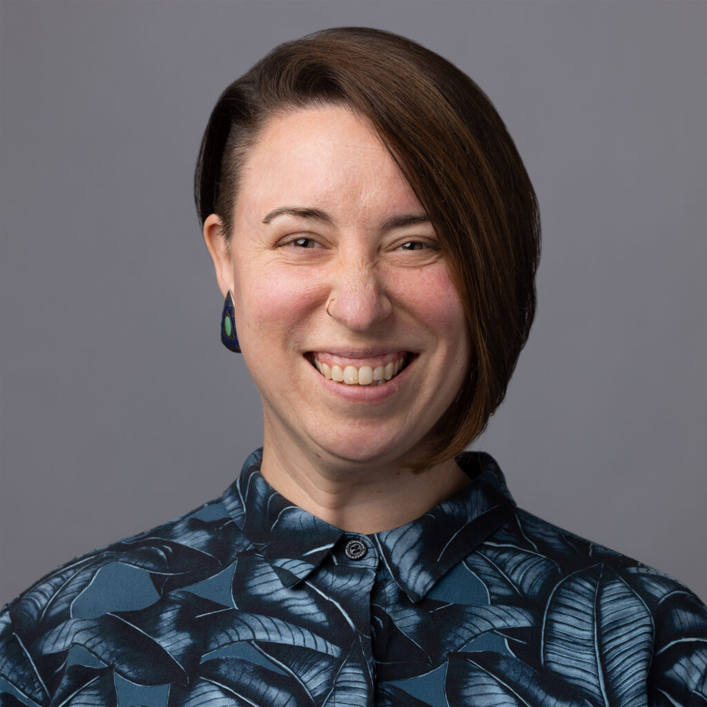 The author, a smiling White woman with short asymmetrical brown hair wearing a blue button down shirt and vintage earrings